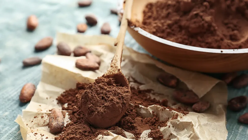 Wooden spoon heaped with unsweetened cocoa powder on parchment paper with scattered cocoa beans
